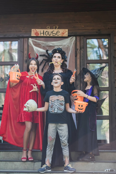 spooky family in halloween costumes holding sweets, buckets and skull on decorated porch - Photo, image