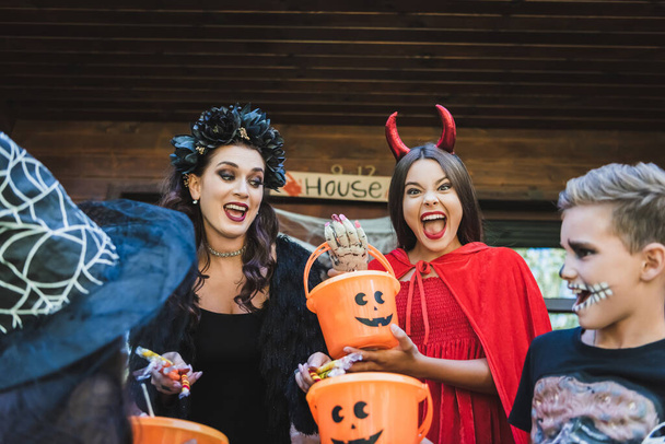 woman with candies near excited kids holding buckets and shouting in creepy halloween costumes - Foto, Imagen