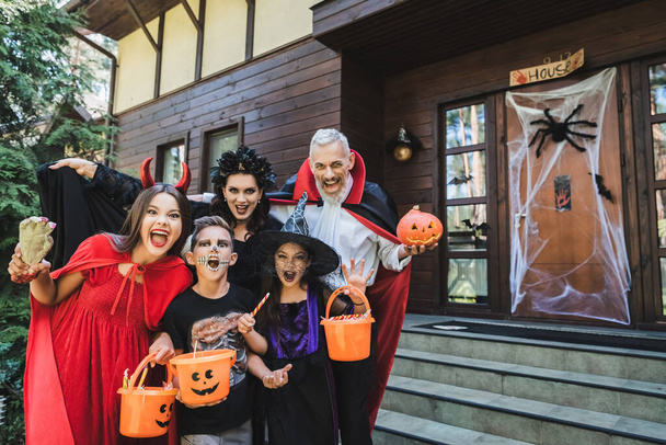 family in creepy costumes holding halloween attributes and shouting near house - Foto, Bild
