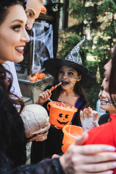 счастливые дети в жутких костюмах на Хэллоуин едят конфеты рядом с размытыми родителями - Фото, изображение