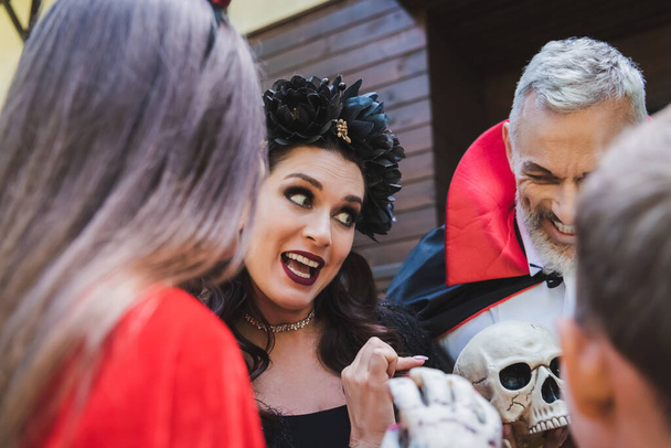 Frau im Halloween-Kostüm zeigt auf Totenkopf, während sie mit verschwommenen Kindern spricht - Foto, Bild
