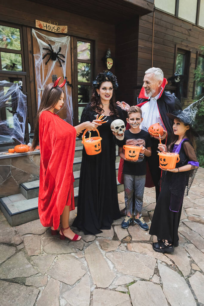cheerful family in spooky costumes holding buckets of sweets near cottage with halloween decoration - Fotó, kép