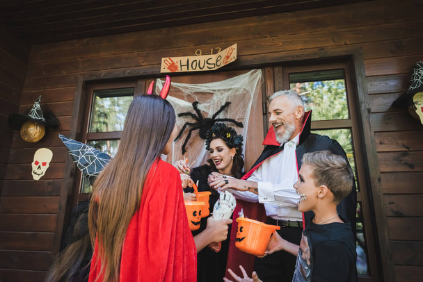 excited kids in halloween costumes holding buckets with candies near neighbors on porch - Foto, afbeelding