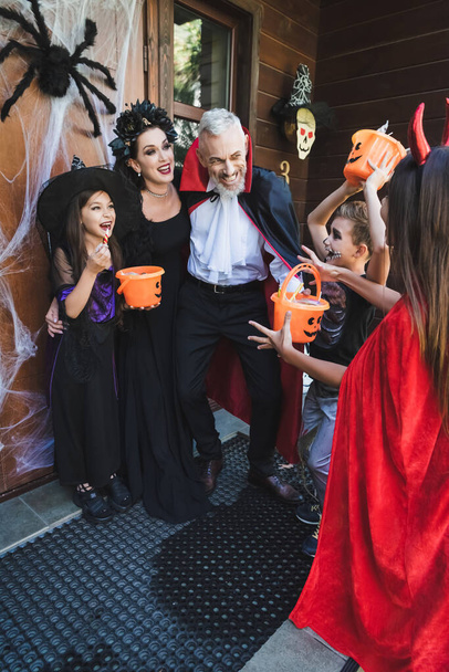 família feliz em trajes assustadores segurando baldes com doces no alpendre com decoração halloween - Foto, Imagem