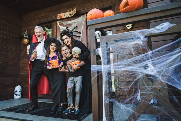 familia alegre en trajes espeluznantes riendo en el porche con decoración de halloween - Foto, Imagen