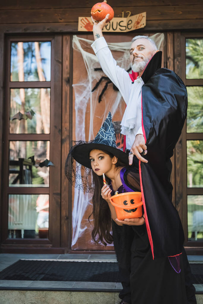 homme en costume de vampire halloween tenant citrouille sculptée dans la main levée près de la fille en chapeau de sorcière  - Photo, image