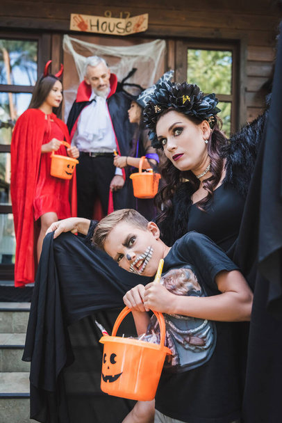 Moeder en zoon in griezelige halloween kostuums op zoek naar camera in de buurt van familie op wazig veranda - Foto, afbeelding
