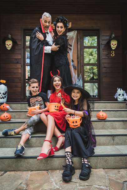parents in halloween costumes looking at kids sitting on cottage stairs with buckets of sweets - Fotoğraf, Görsel