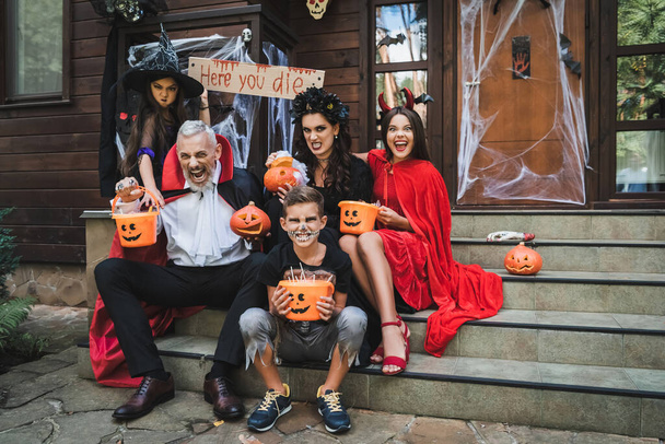 ouders en kinderen in griezelige halloween kostuums grimmig terwijl zitten op cottage trappen - Foto, afbeelding