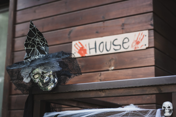 Gruseliger Totenkopf mit Hexenhut und schwarzem Schleier neben verschwommener Karte mit Hausschrift und blutigen Handabdrücken - Foto, Bild