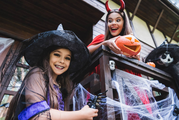 low angle view of cheerful sisters in halloween costumes playing on porch with decoration - Zdjęcie, obraz