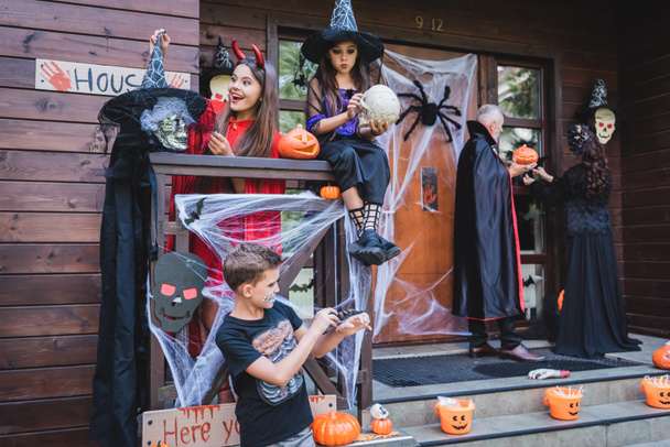family in halloween costumes decorating porch of wooden cottage with scary attributes - Fotoğraf, Görsel