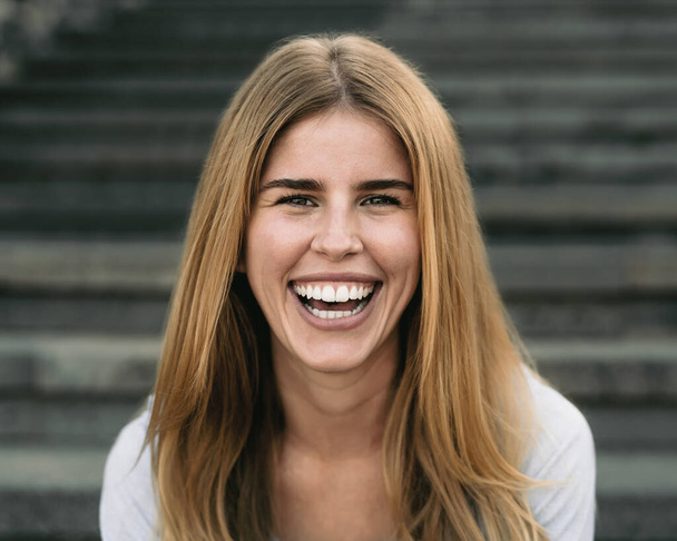 Ευτυχισμένη νεαρή γυναίκα χαμογελά στην κάμερα, ενώ κάθεται σε αστικές σκάλες - Φωτογραφία, εικόνα