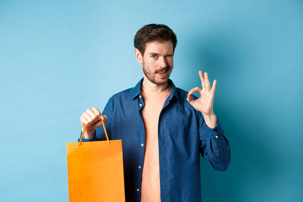 Moderni mies rento asu näyttää ostoskassi ja kunnossa merkki, silmää kameralle, suosittelee kauppa, seisoo sinisellä taustalla - Valokuva, kuva