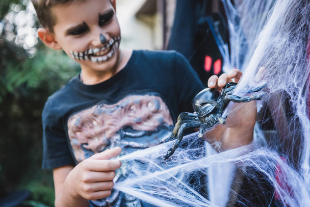 chico borroso en disfraz de esqueleto de Halloween jugando con araña de juguete cerca de la red de araña decorativa - Foto, imagen
