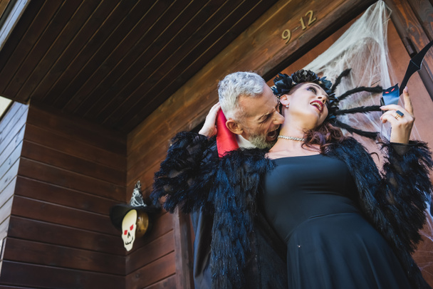 low angle view of man in halloween vampire costume biting neck of wife on cottage porch - Photo, Image