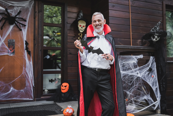 щасливий зрілий чоловік у костюмі вампіра Хеллоуїн стоїть на прикрашеному ганку з паперовим вирізом кажана
 - Фото, зображення