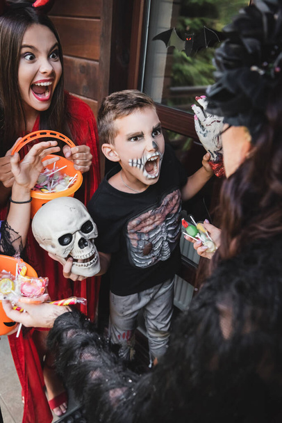 blurred woman holding candies near spooky kids in halloween costumes growling while holding buckets - Foto, imagen
