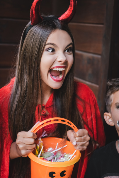 girl in devil halloween costume holding bucket with sweets and screaming near brother - Zdjęcie, obraz