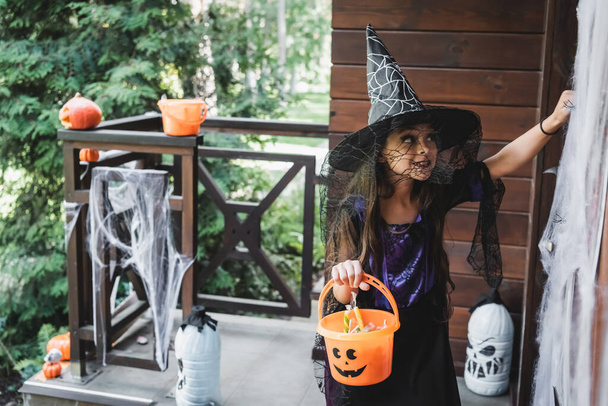 niño en traje de Halloween bruja sosteniendo cubo con dulces mientras llama a la puerta en el porche decorado - Foto, Imagen