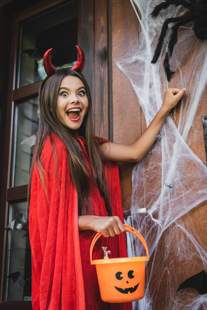 excited girl in devil halloween costume holding bucket while knocking at door with spider net - 写真・画像