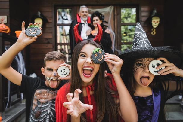 spooky kids grimacing holding halloween cookies and grimacing near blurred parents  - Foto, imagen