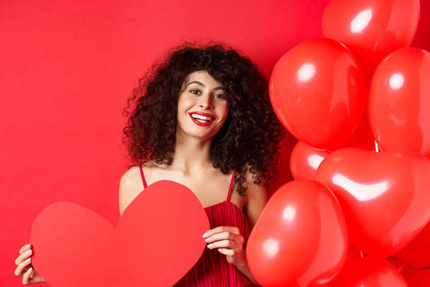 Happy valentines day. Romantic girl in dress waiting for true love, showing big red heart and standing near balloons on red background - Fotó, kép