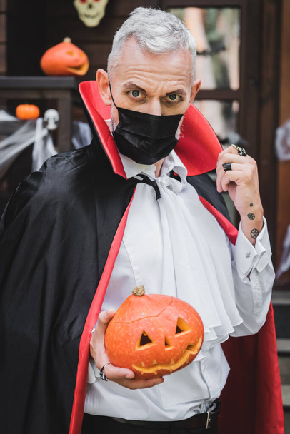 middle aged man in black medical mask and vampire halloween costume holding carved pumpkin while looking at camera  - 写真・画像