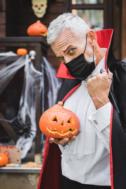 hombre maduro en traje de Halloween vampiro y máscara médica negro mirando a la cámara mientras sostiene calabaza tallada - Foto, imagen