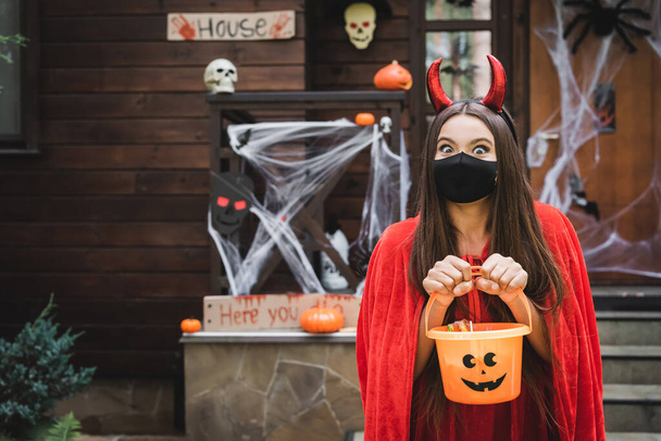 girl in devil costume and black medical mask standing with bucket of sweets near blurred cottage with halloween decoration - Fotó, kép