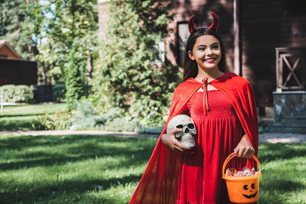 happy child in red demon costume standing with skull and bucket of candies outdoors - Фото, зображення