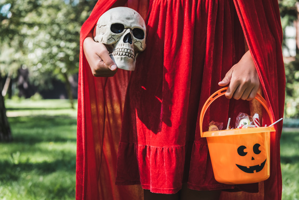 partial view of girl wearing red halloween costume and holding skull and bucket of candies - Zdjęcie, obraz