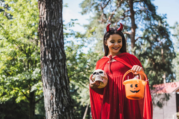 hymyilevä lapsi paholainen halloween puku pitämällä kallo ja ämpäri karkkeja metsässä - Valokuva, kuva