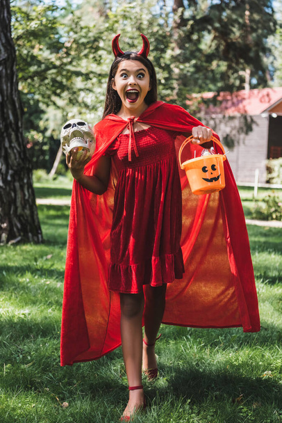astonished girl in demon halloween costume shouting while standing with skull and bucket of candies outdoors - Photo, Image