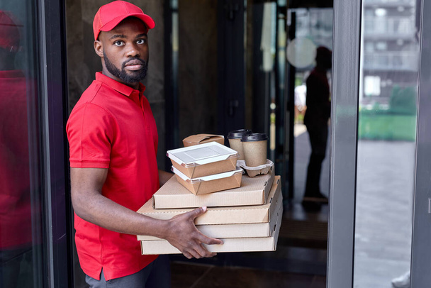 Food delivery service by african courier. Delivery man in red uniform with pizza boxes - Foto, imagen