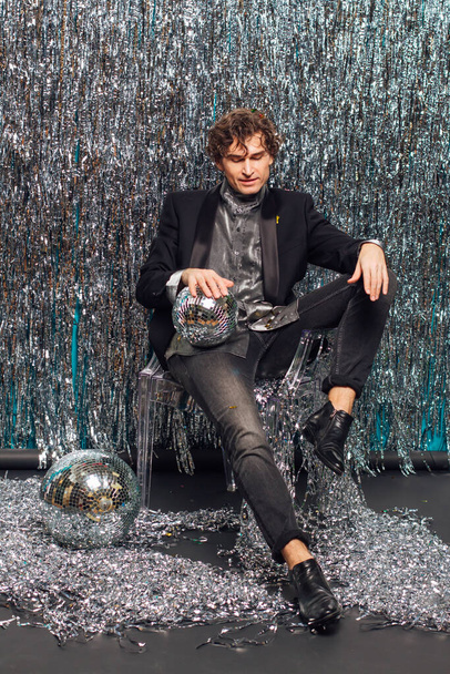 Tall attractive man sitting on the floor over the sparkle background and mirror disco ball holding glass of champagne. Party concept. - Zdjęcie, obraz