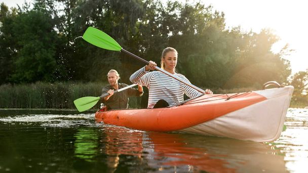 Motivated young couple of friends boating together on river with trees in the background - 写真・画像