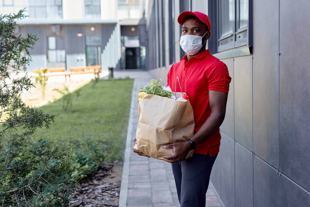 Handsome african delivery worker man in cap and medical mask carrying packet with fresh food - Фото, изображение