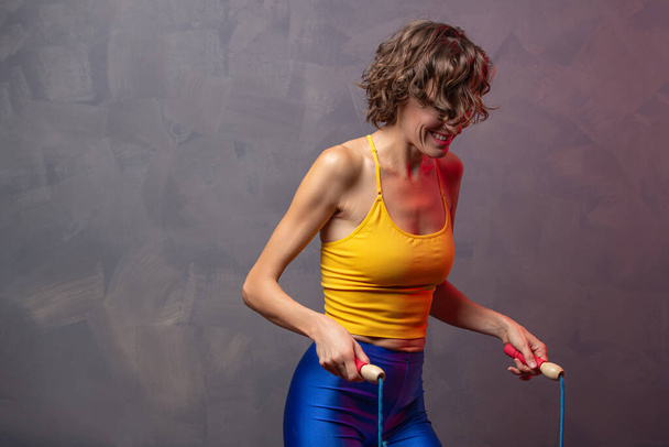 Happy and young caucasian woman with perfect body skipping rope while exercising in studio against grey background - Photo, Image