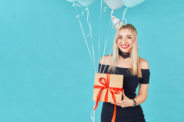 Beautiful smiling woman in black dress and birthday hat holding gift box and pastel air balloons against blue background. Happy young girl celebrating birthday party. - Photo, image