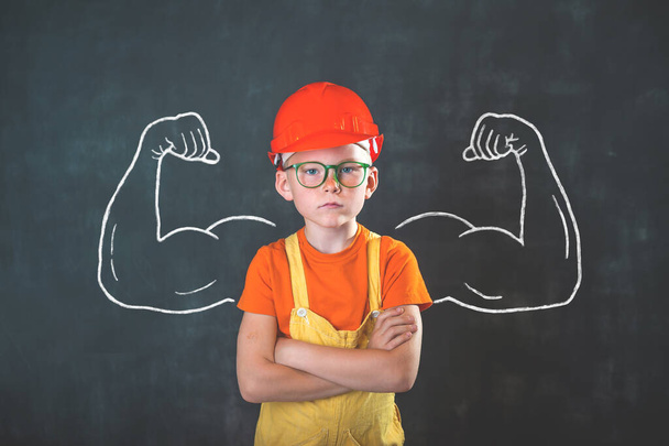 Portrait of smart child in safety red helmet and builder uniform with big muscles. A school boy dreams of becoming a builder guy. Children profession. Smart kid. Confidentconcept - Photo, Image