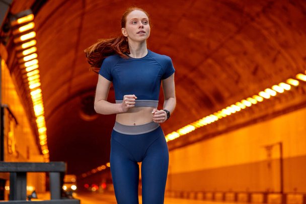 portrait of young red-haired lady running forward, exercising and training alone along road - Photo, Image