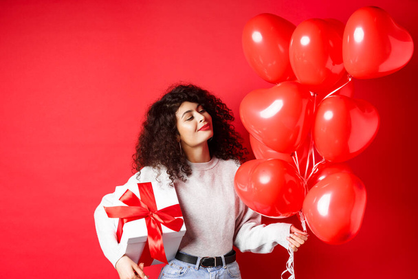 Ρομαντικό γυναίκα αναζητούν ονειρική σε μπαλόνια καρδιά από τον εραστή, κρατώντας δώρο Ημέρα του Αγίου Βαλεντίνου σε χαριτωμένο τυλιγμένο κουτί, στέκεται σε κόκκινο φόντο - Φωτογραφία, εικόνα