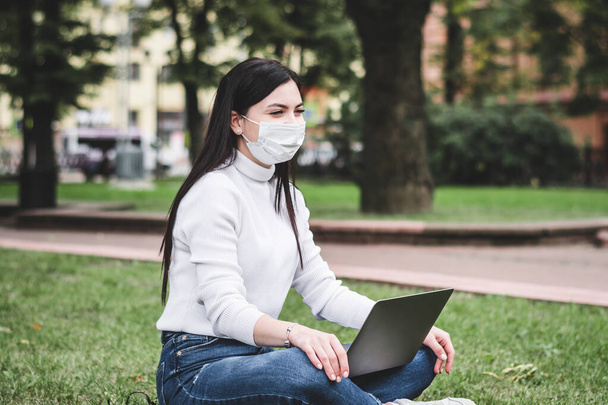 Młoda dziewczyna w masce ochronnej pracuje w parku na laptopie i z telefonem w rękach - Zdjęcie, obraz