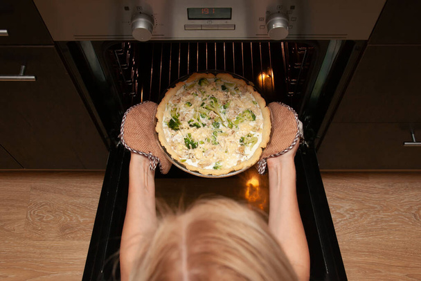 Vrouw neemt een open shortbread taart met kip en broccoli, bestrooid met kaas uit de oven in haar keuken. - Foto, afbeelding