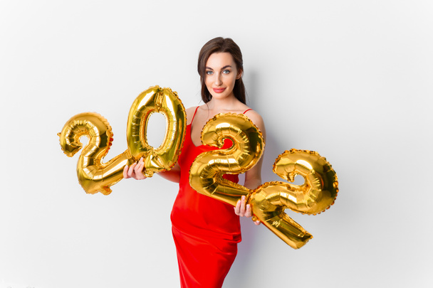 Young smiling woman in red cocktail dress with bright make-up celebrating New Year 2022 and holding golden balloons 2022 in hands on white background - Foto, immagini