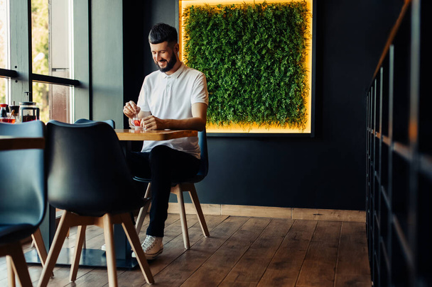 Sonriente joven atractivo hombre disfrutando de té de frutas en la cafetería mientras está sentado en la mesa en la cafetería moderna, hombre sosteniendo la taza y sonriendo mientras está sentado en la mesa - Foto, Imagen