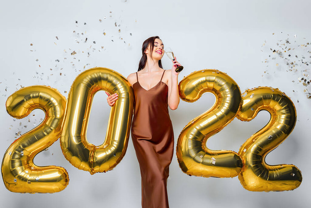 Happy young brunette woman with glass of champagne near golden 2022 balloons on white background. New Year celebration. Wearing fashionable glamor dress. Confetti background. Drinking champagne - Fotoğraf, Görsel