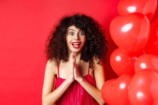 Valentinstag. Aufgeregte kaukasische Frau im Kleid springt vor Staunen, schaut etwas höhnisch an, will ein Produkt bekommen, steht in der Nähe von Herzballons, weißer Hintergrund - Foto, Bild
