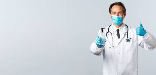 Covid-19, prevención de virus, trabajadores sanitarios y concepto de vacunación. Médico satisfecho en máscara médica y guantes thumb-up en la aprobación, recomiendan desinfectante de manos, fondo blanco
 - Foto, imagen